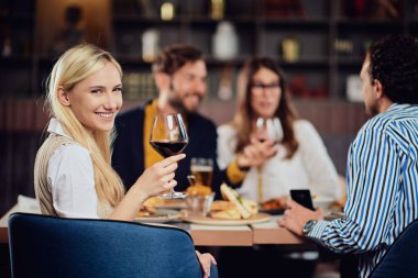 Restoran ve kırmızı şarap tutarak cam oturan güzel Kafkas blonede kadın portresi. Arka planda arkadaşlar akşam yemeği.
