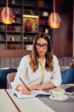 Young beautiful serious Caucasian brunette sitting at cafe and writing in agenda tasks. Remote business concept.