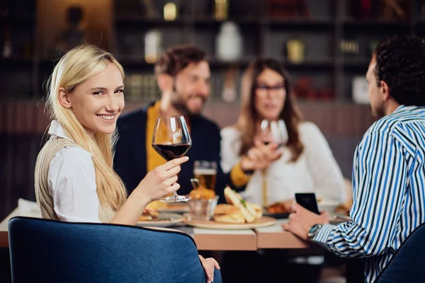 Portré gyönyörű kaukázusi blonede nő ül az étteremben és a gazdaság pohár vörösbort. Háttérben barátai vacsora. — Stock Fotó