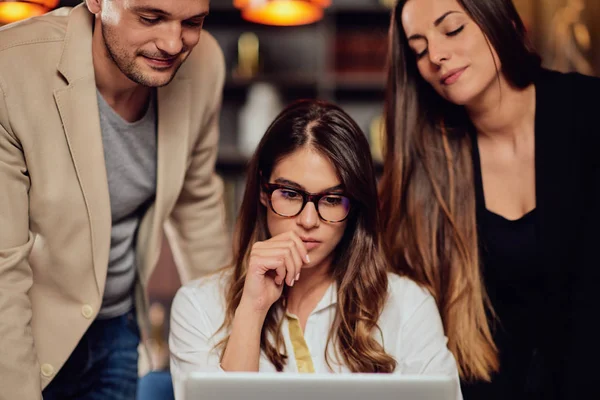 Charming buisnesswoman with brown hair and dressed elegant sitting at restaurant and looking at laptop. Next to her standing two colleagues and looking at laptop, too. — Stock Photo, Image