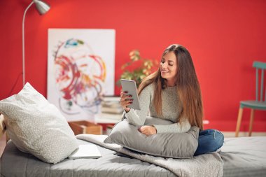 Rahat yatak odasında yatak yastık tur ile oturan ve akıllı telefon kullanarak muhteşem gülümseyen beyaz esmer giyinmiş.