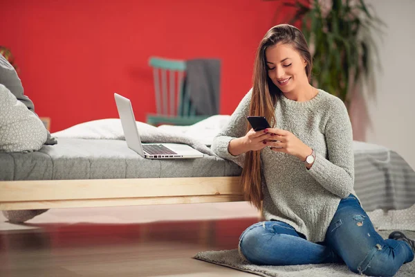 Красивая кавказская брюнетка одета непринужденно, сидя на полу и опираясь на кровать в спальне, разговаривая по телефону. На кровати ноутбук . — стоковое фото