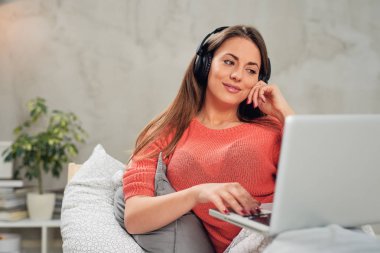 Müzik dinleme ve yatak odasında yatağın üstünde otururken dizüstü kullanarak muhteşem beyaz gülümseyen esmer.