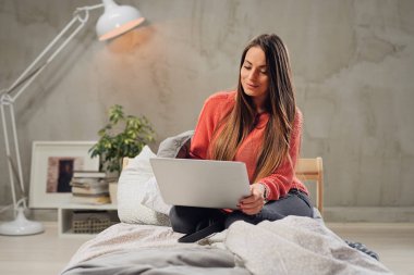 Güzel gülümseyen beyaz esmer yatak odasında yatağın üstüne oturan ve dizüstü bilgisayar kullanarak.