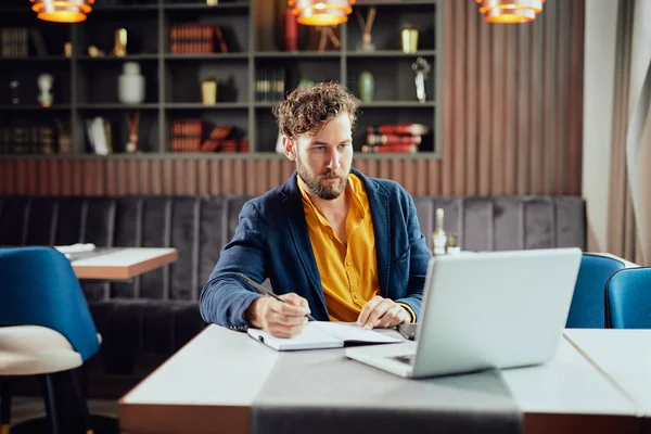 Young serious bearded Caucasian blogger dressed smart casual writing notes in agenda and looking at laptop while sitting in cafeteria. — Stockfoto