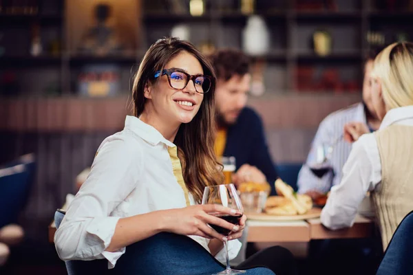 Portrait de magnifique brune regardant la caméra et tenant un verre avec du vin rouge tout en étant assis au restaurant. En arrière-plan amis manger dîner . — Photo