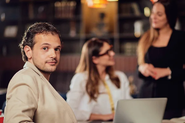 Jonge Arabische zakenman gekleed slim casual zitten in coffeeshop op vergadering en kijken over de schouder op de camera. Op de achtergrond bespreken zijn collega 's over het project. — Stockfoto