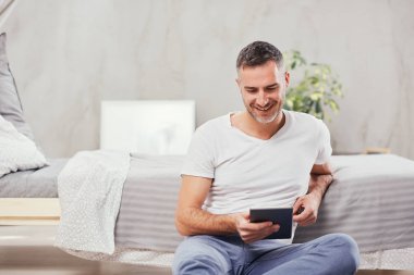 Yakışıklı beyaz orta yaşlı adam yatak odasında yerde oturan ve yatağa yaslanmış. El ele tablet.