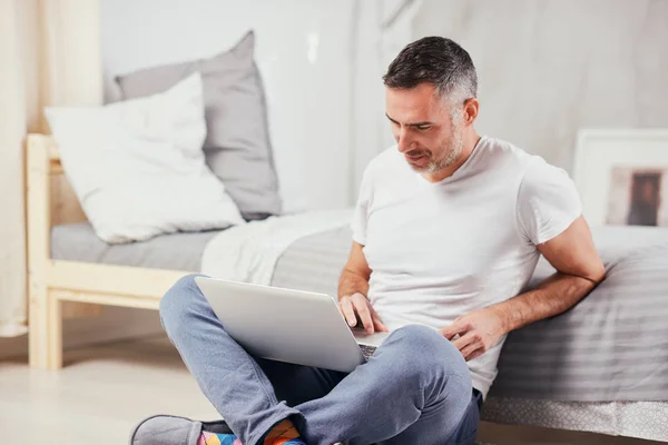 Handsome middle aged man sitting on the floor wirh crossed legs, leaning on bed and using laptop. — Stock Photo, Image