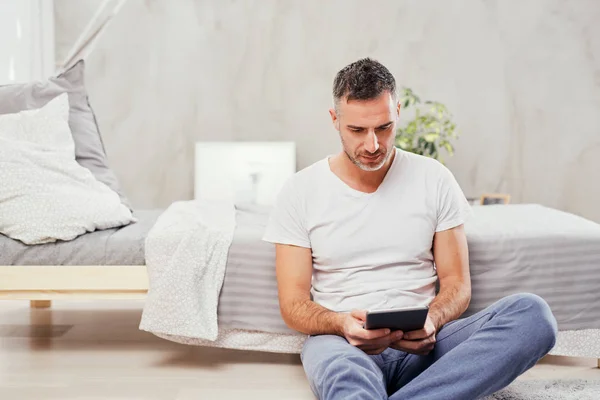 Handsome Caucasian middle-aged man sitting on the floor in bedroom and leaning on bed. In holding hands tablet. — Stock Photo, Image