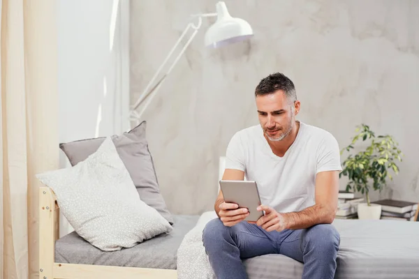 Caucásico hombre de cuarenta años sentado en la cama en el dormitorio y el uso de la tableta . — Foto de Stock