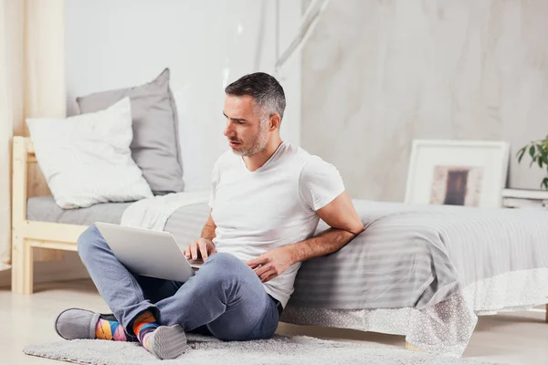 Handsome middle aged man sitting on the floor wirh crossed legs, leaning on bed and using laptop. — Stock Photo, Image