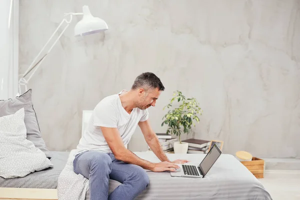 Knappe Kaukasische midden oude mens casual gekleed met behulp van laptop zittend op bed in de slaapkamer. — Stockfoto