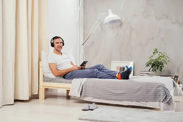 Uomo caucasico di mezza età vestito casual rilassante sul letto in camera da letto, ascoltare musica e utilizzando tablet . — Foto Stock