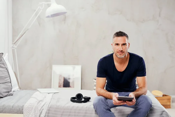 Uomo caucasico sulla quarantina seduto sul letto in camera da letto e utilizzando tablet . — Foto Stock