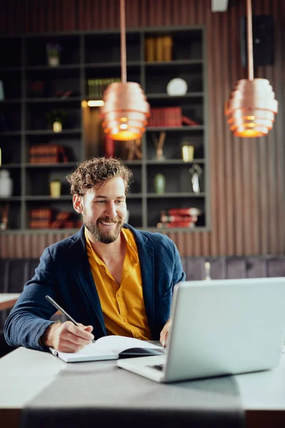 Joven blogger caucásico barbudo sonriente vestido elegante escritura casual notas en la agenda y mirando a la computadora portátil mientras está sentado en la cafetería . — Foto de Stock