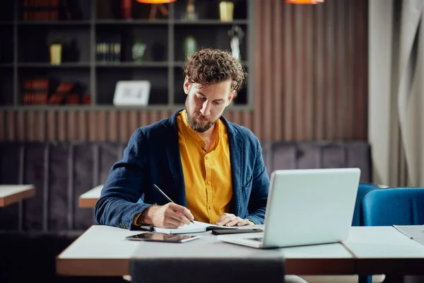 Young serious bearded Caucasian blogger dressed smart casual writing notes in agenda and looking at laptop while sitting in cafeteria. — Stok fotoğraf