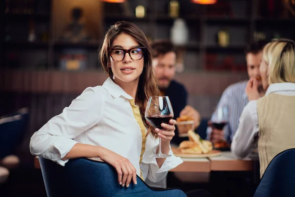 Retrato de linda morena olhando para longe e segurando vidro com vinho tinto enquanto sentado no restaurante. No fundo amigos comendo restaurante . — Fotografia de Stock