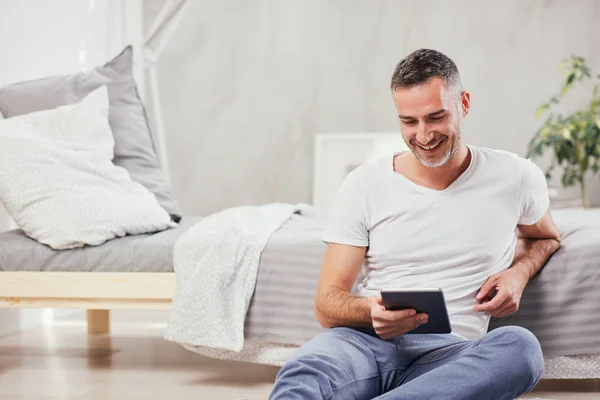 Knappe Kaukasische middelbare leeftijd man zittend op de vloer in de slaapkamer en leunend op bed. In handen tablet. — Stockfoto