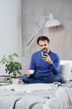 Çekici gülümseyen Arap adam rahat yatak odası, yatakta oturup kahve içme ve akıllı telefon mesaj attığın için kullanarak giyinmiş.