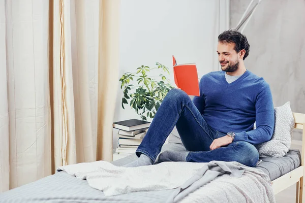 Vrolijke schattig Arabische man gekleed casual boek lezen en ontspannen zittend op het bed in de slaapkamer. — Stockfoto