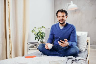 Çekici gülümseyen Arap adam rahat yatak odası, yatakta oturup kahve içme ve akıllı telefon mesaj attığın için kullanarak giyinmiş.