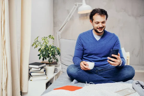 Atractivo hombre árabe sonriente vestido casual sentado en la cama en el dormitorio, beber café y usar el teléfono inteligente para enviar mensajes de texto . — Foto de Stock