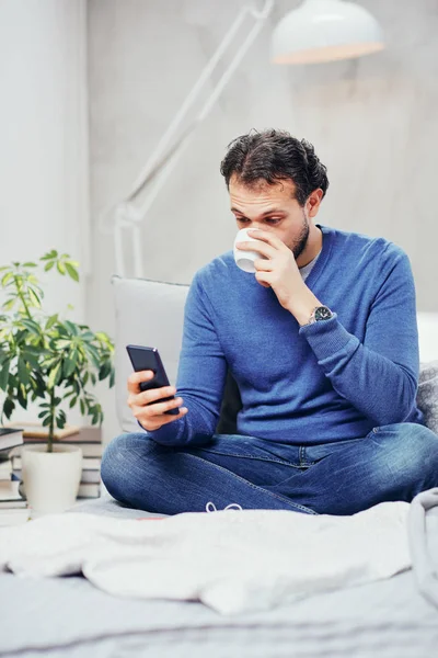 Attractive smiling arab man dressed casual sitting on bed in bedroom, drinking coffee and using smart phone for texting. — Stock Photo, Image