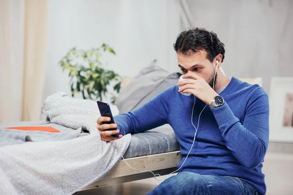 Attractive arab man dressed casual sitting on floor in bedroom, listening music over smart phone and drinking coffee. — Stock Photo, Image