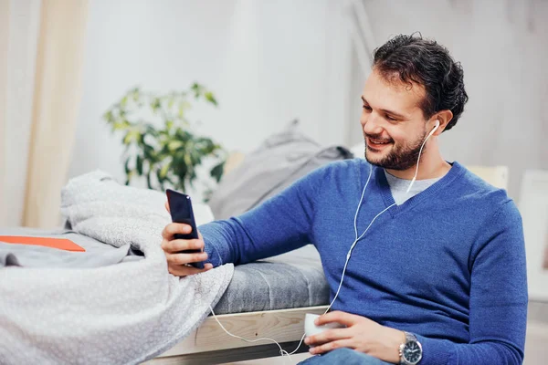 Attractive arab man dressed casual sitting on floor in bedroom, listening music over smart phone and drinking coffee. — Stock Photo, Image