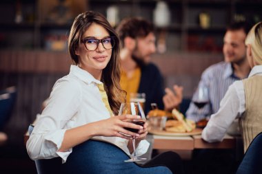 Güzel esmer kamera bakıyor ve Restaurat otururken kırmızı şarap ile cam tutarak portresi. Arka planda arkadaşlar yeme Diner.