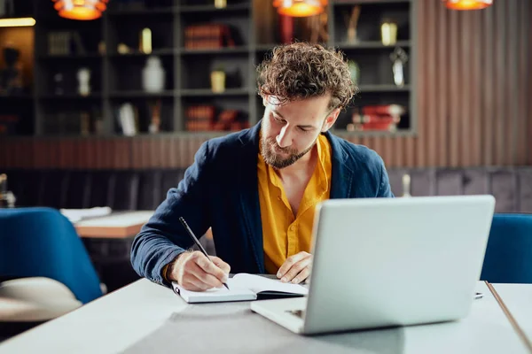 Unga allvarliga skäggiga kaukasiska bloggare klädd smart casual skriva anteckningar i agenda medan du sitter i cafeteria. — Stockfoto