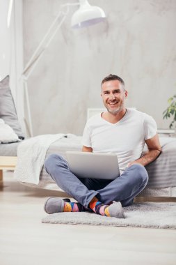 Kat wirh üzerinde oturan yakışıklı orta yaşlı adam yatağa yaslanmış ve dizüstü bilgisayar kullanarak bacak, bacak.