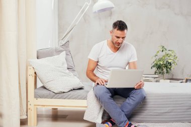 Yakışıklı beyaz orta yaşlı adam rahat giyinmiş yatak yatak odasında otururken dizüstü bilgisayar kullanarak.