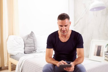 Beyaz adam yatakta yatak odasında oturma ve tablet kullanarak kırklı.