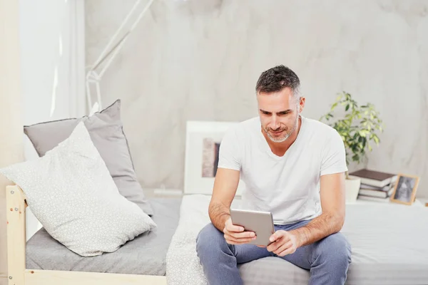 Caucasian man in forties sitting on bed in bedroom and using tablet. — Stock Photo, Image