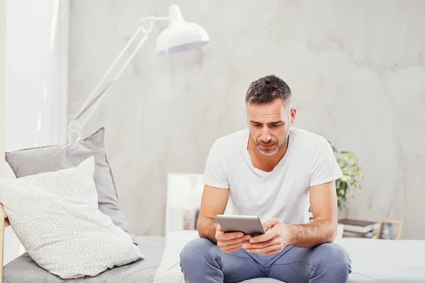 Caucasian man in forties sitting on bed in bedroom and using tablet. — Stock Photo, Image
