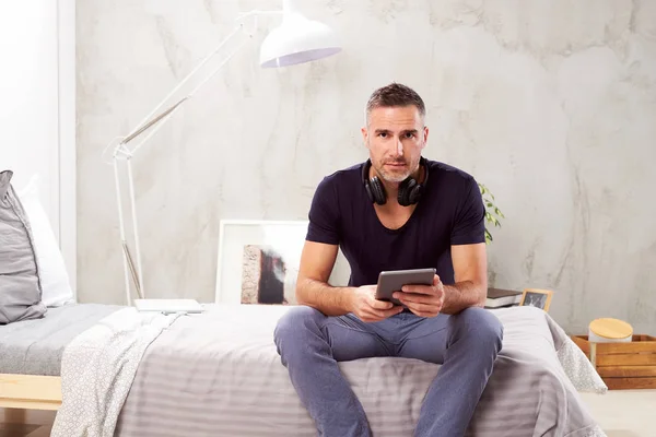 Uomo caucasico sulla quarantina seduto sul letto in camera da letto e utilizzando tablet . — Foto Stock