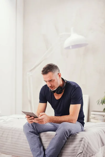 Caucasian man in forties sitting on bed in bedroom and using tablet. — Stock Photo, Image
