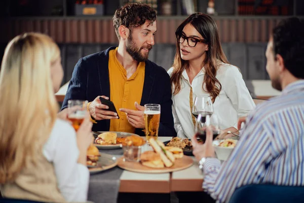 Молодая привлекательная белая пара сидит в ресторане и смотрит на смартфон. На переднем плане сидят их друзья и пьют. . — стоковое фото