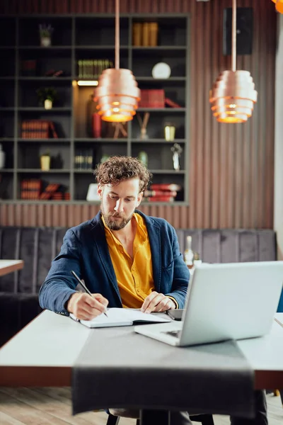 Young serious bearded Caucasian blogger dressed smart casual writing notes in agenda and looking at laptop while sitting in cafeteria. — Stok fotoğraf