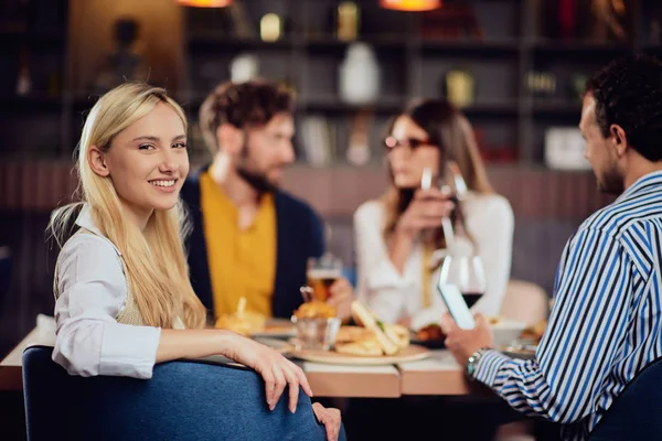 Jovem sorridente caucasiano mulher bonita loira vestida inteligente casual sentado à mesa no restaurante e olhando por cima do ombro. No fundo, seus amigos estão jantando . — Fotografia de Stock