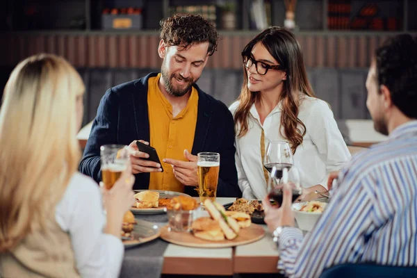 Mladý atraktivní kavkazský pár sedí v restauraci a dívá se na chytrý telefon. V popředí jejich přátelé sedí a popíjejí. — Stock fotografie