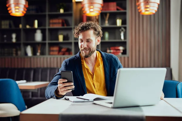 Unga kaukasiska affärsman klädd smart casual med hjälp av smart telefon när du sitter i kaféet. På skrivbordet laptop. — Stockfoto