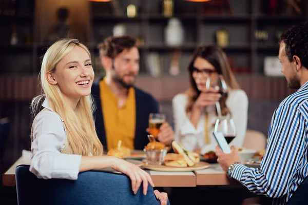 Jovem sorridente caucasiano mulher bonita loira vestida inteligente casual sentado à mesa no restaurante e olhando por cima do ombro. No fundo, seus amigos estão jantando . — Fotografia de Stock