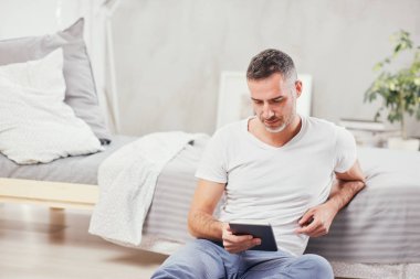 Yakışıklı beyaz orta yaşlı adam yatak odasında yerde oturan ve yatağa yaslanmış. El ele tablet.