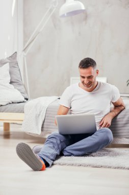 Yakışıklı orta yaşlı adam yerde oturan, yatağa yaslanmış ve dizüstü bilgisayar kullanarak.