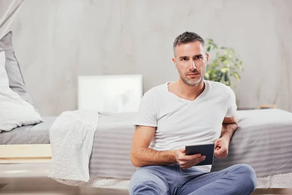 Guapo hombre caucásico de mediana edad sentado en el suelo en el dormitorio y apoyado en la cama. En la tableta de la mano . — Foto de Stock