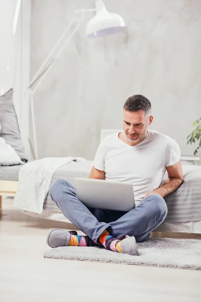 Knappe middelste leeftijd man zittend op de vloer, leunend op bed en met behulp van laptop. — Stockfoto