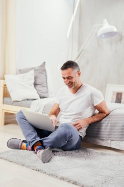 Yakışıklı orta yaşlı adam yerde oturan, yatağa yaslanmış ve dizüstü bilgisayar kullanarak.